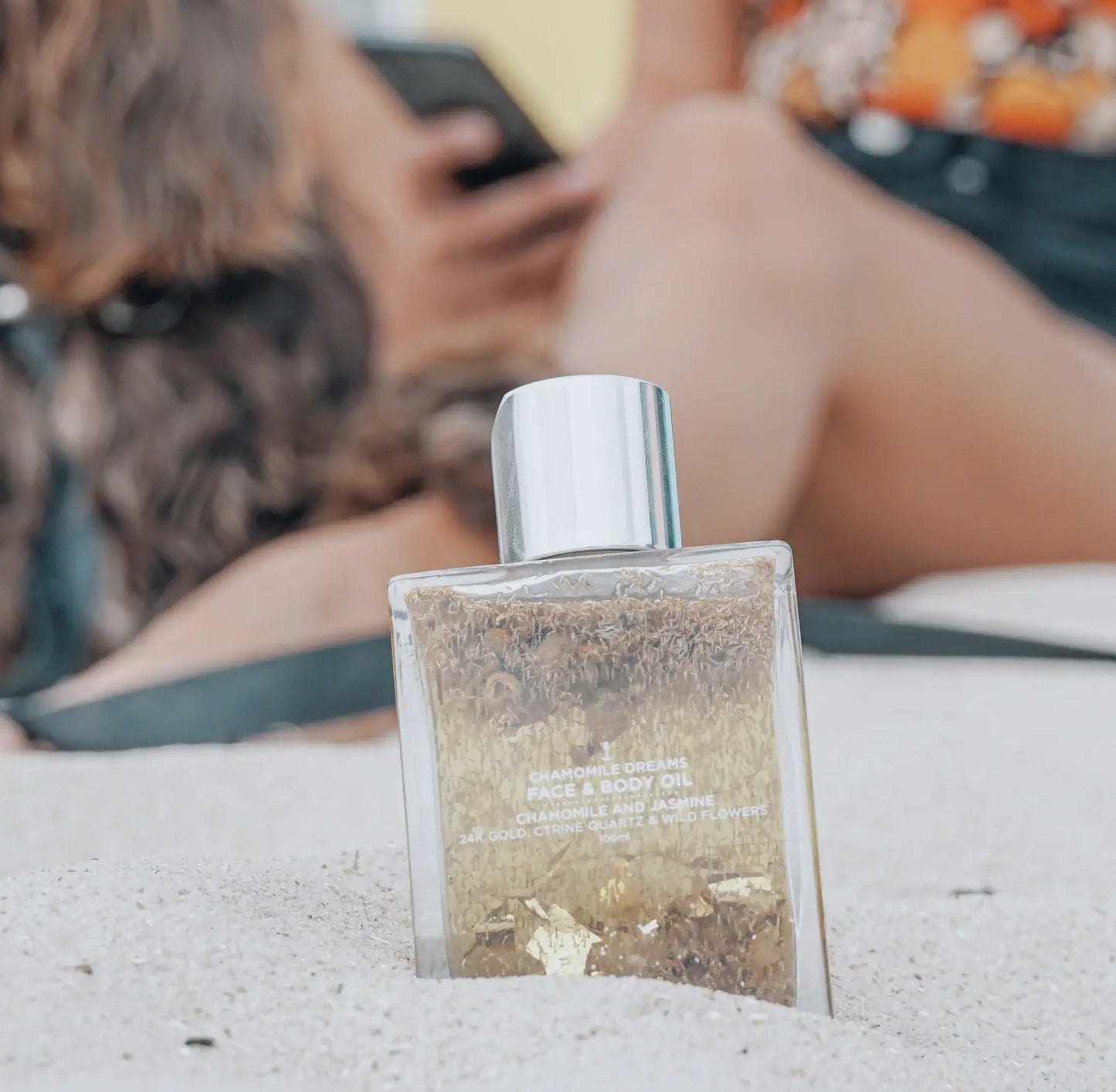 Chamomile Dreams Face & Body Oil - 100ml bottle, featuring a soft, calming design with a subtle floral pattern, resting on a white sand surrounded by fresh chamomile flowers. The oil promotes relaxation and nourishment for both face and body.